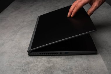 Erkek el, gri bir masada parlak siyah bir dizüstü bilgisayarı kapatıyor, işi tamamlamayı, verimliliği veya teknoloji kullanımını sembolize ediyor.