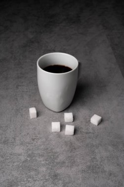 Gri bir yüzey üzerinde şeker küpleriyle çevrili siyah kahve dolu beyaz bir kupa..