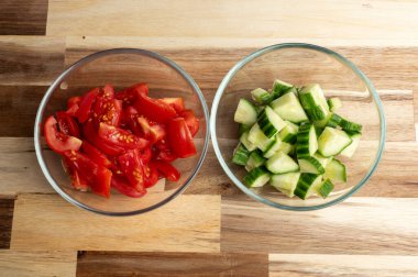 İki cam kase içinde doğranmış sebzeler var: biri doğranmış domatesli, diğeri de dilimlenmiş salatalıklı, tahta bir yüzeye yerleştirilmiş.