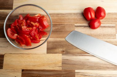 Tahta bir yüzey üzerinde bir kase doğranmış domates, yanında da tüm domatesler ve bir bıçak.