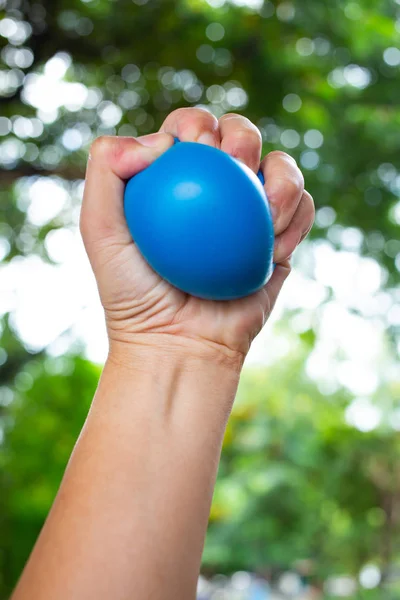 La main gauche de la femme serrant la balle de stress bleue sur fond de jardin vert bokeh, concept d'exercice et de massage — Photo
