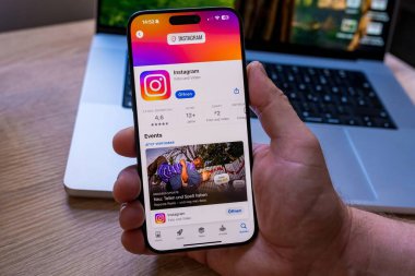 App Store 'da Instagram uygulama sayfasını gösteren akıllı telefonu tutan bir elin yakın çekimi. Ekranda uygulama derecelendirmesi, yükleme düğmesi ve resimlerin önizlemesi, sosyal medya kullanımı, mobil teknoloji ve dijital iletişim sembolize edilir. Nuremberg