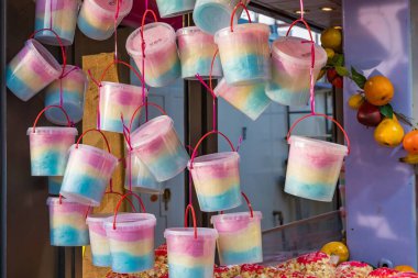 Şeffaf plastik kovalara doldurulmuş renkli pamuk şekerler adil bir ahırda asılı duruyor. Pastel pembe, mavi ve sarı şeker karnavallarda, lunaparklarda ve festivallerde, neşeyi, çocukluğu ve kutlamaları sembolize eden popüler bir tatlı ikramdır..