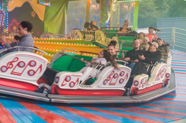 Müzik Ekspresi 'nin keyfini çıkaran bir grup insan bir karnavalda geziyor. Renkli ışıklar ve hızlı hareketle aydınlatılmış panayır cazibesi heyecan, neşe ve geleneksel eğlence eğlencesini sembolize eden canlı bir atmosfer yaratır.