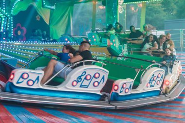 Bir lunaparkta Music Express 'e binen insanların yakın çekimi. Parlak ışıklar ve hızlı hareketle aydınlatılmış yolculuk heyecan, neşe ve geleneksel fuar alanını sembolize eden dinamik bir karnaval atmosferi yaratır.