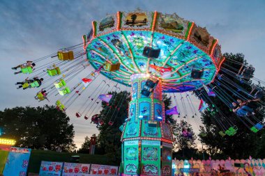 Illuminated swing ride carousel chair-o-planes operating at a fairground during the evening. People enjoy the ride as colorful lights shine brightly against the evening sky, capturing the festive atmosphere of a carnival or amusement park