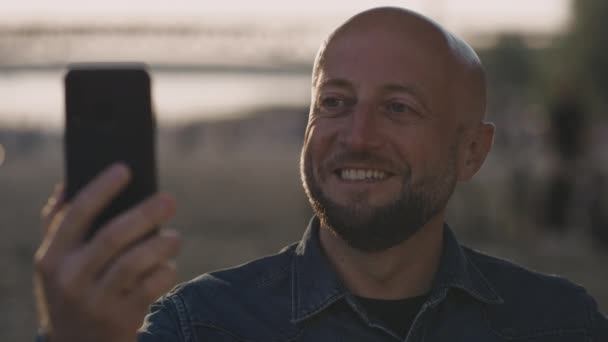 Glücklicher, glatzköpfiger Mittvierziger mit stylischem Bart steht bei Sonnenuntergang am Strand. Er hat ein nettes Videotelefonat auf seinem Handy mit jemandem, den er mag. Er lächelt wild. — Stockvideo