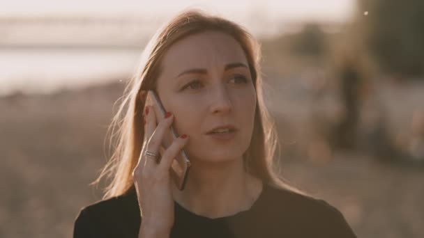 Egy fiatal nő beszél a telefonban. A tengerparton áll egy gyönyörű naplementében.. — Stock videók