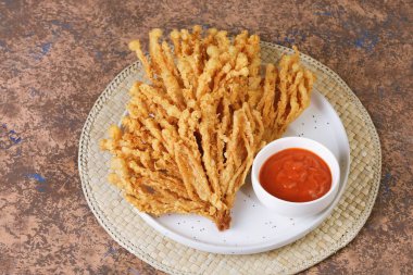 Golden brown crispy fried enoki mushroom snack served with chili sauce on white plate. Popular Asian street food snack.