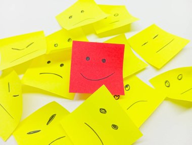 Mutlu yüzlü tek bir kırmızı yapışkan not hüzünlü veya nötr yüzlü, bireyselliği ve olumlu bir görünümü sembolize eden sarı yapışkan notlar yığınından çıkar..