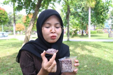 A candid shot of a young Indonesian woman in a black hijab and glasses, looking at a chocolate-covered wafer with a funny, puckered expression on her face.