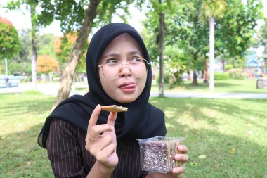 A fun, candid portrait of a young Indonesian woman in a black hijab, wearing glasses, holding a chocolate wafer and sticking her tongue out playfully.