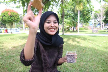 An Indonesian woman in a black hijab smiles while holding up a snack in a park.