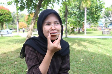 A happy Indonesian woman in a black hijab and glasses bites into a chocolate-covered wafer while sitting on the grass in a park.