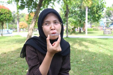 A close-up of a young Indonesian woman in a black hijab and glasses, looking to the side with a puckered expression as she eats a chocolate wafer in a park.
