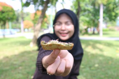 A blurry portrait of an Indonesian woman in a black hijab holding a chocolate-covered wafer towards the camera, with the wafer in sharp focus.