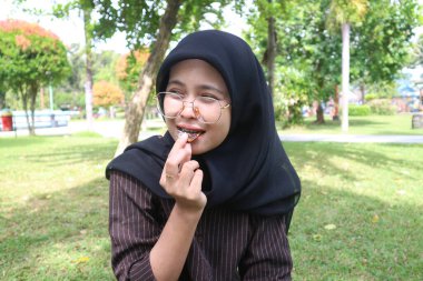 A joyful Indonesian woman in a black hijab smiles and looks away while eating a chocolate wafer in a lush green park.