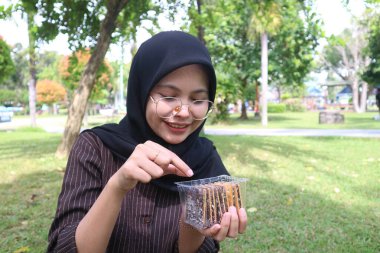 A joyful young Indonesian woman in a black hijab smiles while looking down at a container of chocolate wafers and pointing at them with her finger in a park.