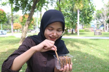 A smiling Indonesian woman in a black hijab and glasses happily selects a chocolate-covered wafer from a plastic container while sitting on the grass in a park.