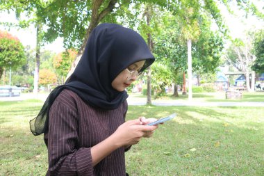 A young woman wearing a black hijab and glasses sits on a grassy lawn in a park, intently looking at and using her smartphone.