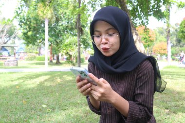 A young woman wearing a black hijab and glasses sits on a grassy lawn in a park, holding her smartphone with a pursed-lip expression. She is concentrating on the phone