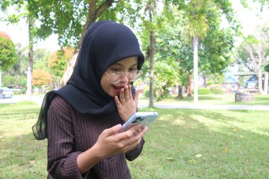 A young woman wearing a black hijab and glasses sits on a grassy lawn in a park, holding her smartphone with one hand over her mouth in an expression of surprise or excitement.