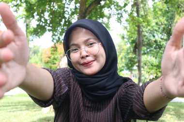 A young woman wearing a black hijab and glasses smiles warmly, reaching her hands out towards the camera in a welcoming gesture, as if taking a selfie or a POV shot.