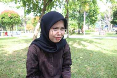 A young woman wearing a black hijab and glasses sits on a grassy lawn in a park, looking to the side with a serious or concerned expression.