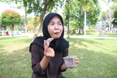 A young woman with a black hijab and glasses sits on a lawn, playfully making a face while eating a chocolate brownie from a plastic container. The background is a blurred park scene with trees.