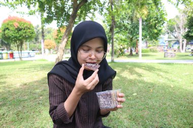 A young woman wearing a black hijab and glasses sits on a grassy lawn in a park, smiling with her eyes closed as she takes a bite of a brownie. She holds a plastic container of brownies in her other hand.