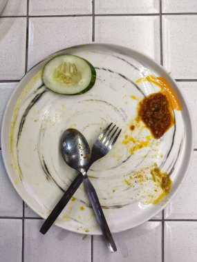 İçinde salatalık dilimi ve artık sosu olan boş beyaz bir tabak, bir kaşık ve çatal eşliğinde fayanslı bir yüzey üzerinde temiz, minimalist ve yemek ve restoran pazarlaması için ideal bir yemek sahnesi yaratıyor..