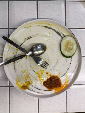 Bir kaşık ve çatallı temiz beyaz tabak, bir salatalık dilimi ve beyaz fayanslı yüzeyde arta kalan baharatlı sos yemek ve restoran pazarlaması için uygun minimalist ve sıradan bir yemek sahnesi yaratıyor..