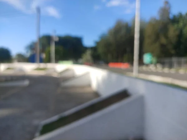 Blurred outdoor scene with bright sunlight, white concrete walls, and a road with green trees in the background