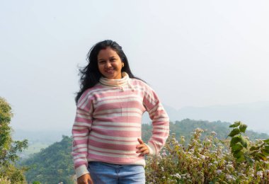 Dağ zirvesinde yeşil çimlerle fotoğraf çeken Hintli kız kameraya bakıyor. Ormanda sportif genç bir kadınla manzara. Seyahat, turizm konsepti.