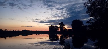 Twilight lake reflecting silhouetted trees under a warm orange-to-blue sky.