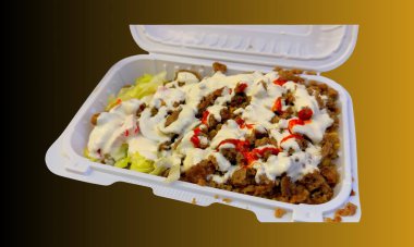 Close up of lamb over rice served in a white takeout container, topped with creamy white sauce and spicy red sauce, accompanied by fresh lettuce salad.