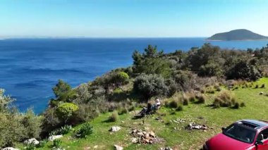 Aerial video of the coast near Kalkan, Turkey. Two cars parked on a grassy hilltop by the sea, with people enjoying a picnic overlooking the vibrant blue Mediterranean and mountains on the horizon. Perfect for travel, road trip, and outdoor. 60fps