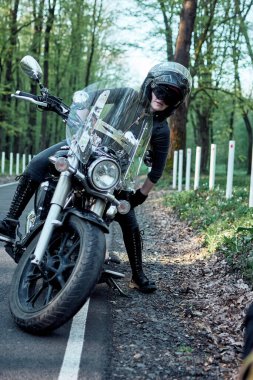 Havalı bir bayan motosikletçi yol kenarında motosikletinin yanında poz verirken görülüyor, kaskı ve şık siyah giysileri var, motosiklet tutkusu ve açık yolu temsil ediyor..