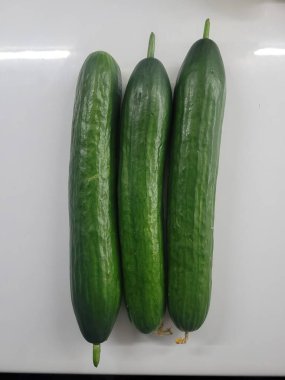 Parlak beyaz bir yüzeye sergilenen üç taze salatalık yığını, dokusunu ve rengini vurgulayarak, mutfak, sağlıklı beslenme veya tarım temalı projeler için uygundur..