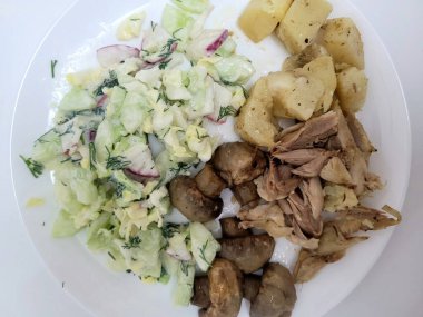 Üzerinde salatalık, turp salatası, salatalık, ekşi krema sosu, patates, mantar ve çeşitli ve sağlıklı bir yemek sergilenen tavuk eti olan beyaz bir tabağın üstünden..