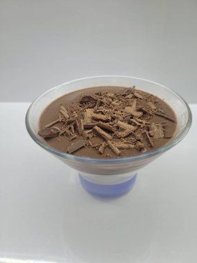 Şeffaf cam bir kasede çikolatalı puding. Çikolata parçacıklarıyla süslenmiş. Tadını çıkarmaya hazır. Beyaz bir arkaplanda çekilen basitlik, tatlının çöküşünü vurguluyor..
