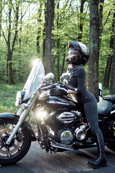 Siyah deri ceketli ve botlu bir kadın ormanda siyah bir motosiklet üzerinde poz veriyor. Ağaçların arasında güneş parlıyor, kendine güven ve güç yayıyor, motosiklet sürücüsünün yaşam tarzını gösteriyor..