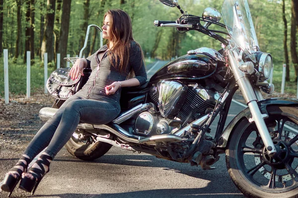 Gri tulumlu esmer bir kadın ve yüksek topuklu ayakkabılar ağaçlarla kaplı bir yolda siyah bir motosiklette poz veriyor. Görüntü, doğal ortamda bir tarz ve özgürlük hissi yakalıyor..