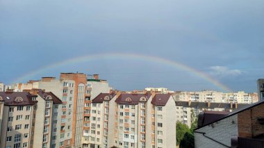 Şehir manzarasında gökkuşağı zarifçe kavis çizer, konut binalarının ve çatıların üstündeki gökyüzü boyunca canlı bir spektrum çizer. .
