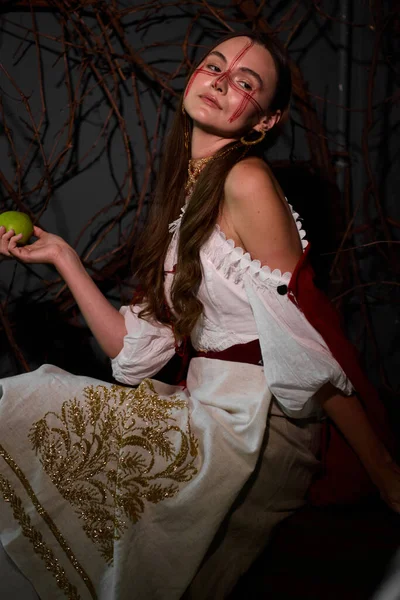 Yüzünde işlemeli, kırmızı makyajlı, beyaz bluzlu genç bir kadın, elinde yeşil bir elma, boynunda ve kulaklarında altın mücevherler, sopalarla dolu bir arka planda oturuyor. .