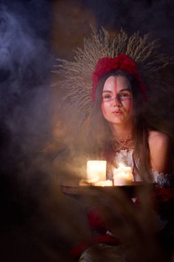 Kızıl savaş boyası ve başlığıyla mum tutan gizemli kız mum ışığında mum yakıyor, gelenek ve ayin atmosferi yaratıyor.. 