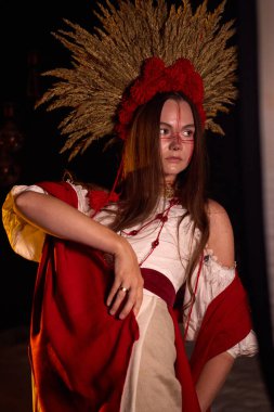 Geleneksel Ukrayna giysisi ve başlığıyla süslenmiş, çarpıcı kırmızı yüz boyası ve büyüleyici bir atmosfer yaratan koyu bir arka plana yoğun bir bakışla bakan genç bir kadının büyüleyici portresi. 