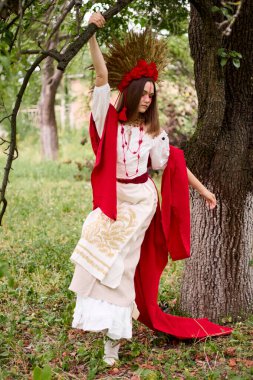 Büyüleyici bir genç kadın geleneksel Ukrayna kıyafetleri içinde, çarpıcı bir buğday çelengi ve kırmızı çiçekli başlıkla süslenmiş, yemyeşil bir ormanda poz veriyor, halk ruhunu ve mirasını somutlaştırıyor. .