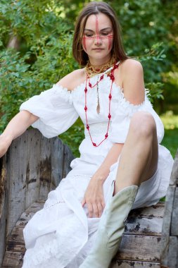 Kırmızı yüz boyası ve mücevheri olan beyaz elbiseli genç bir kadın kırsal bir ahşap yapının üzerine oturmuş çeşitli yaratıcı projeler ve tasarımlar için büyüleyici ve benzersiz bir imaj yaratıyor. 