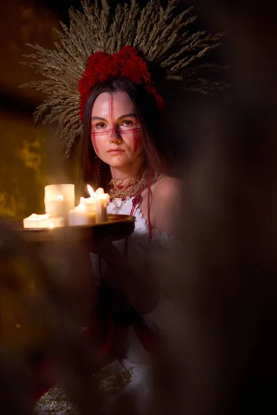 Geleneksel bir elbise ve başlıkla kırmızı yüzlü bir kızın portresi tepside yanan mumlar tutuyor, karanlık bir atmosferde gizem ve ayin çağrıştırıyor. .